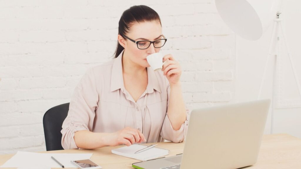 Woman With Glasses Sipping Coffee Cup While Reading On Laptop
